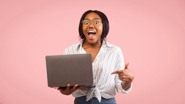 Woman looks extreamly happy as she points to her laptop. Image taken from Adobe Stock